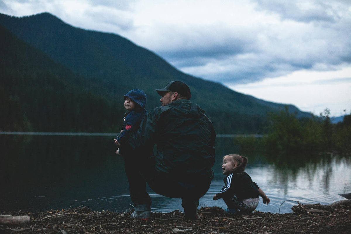 Un adulto y dos niños junto a un lago entre montañas