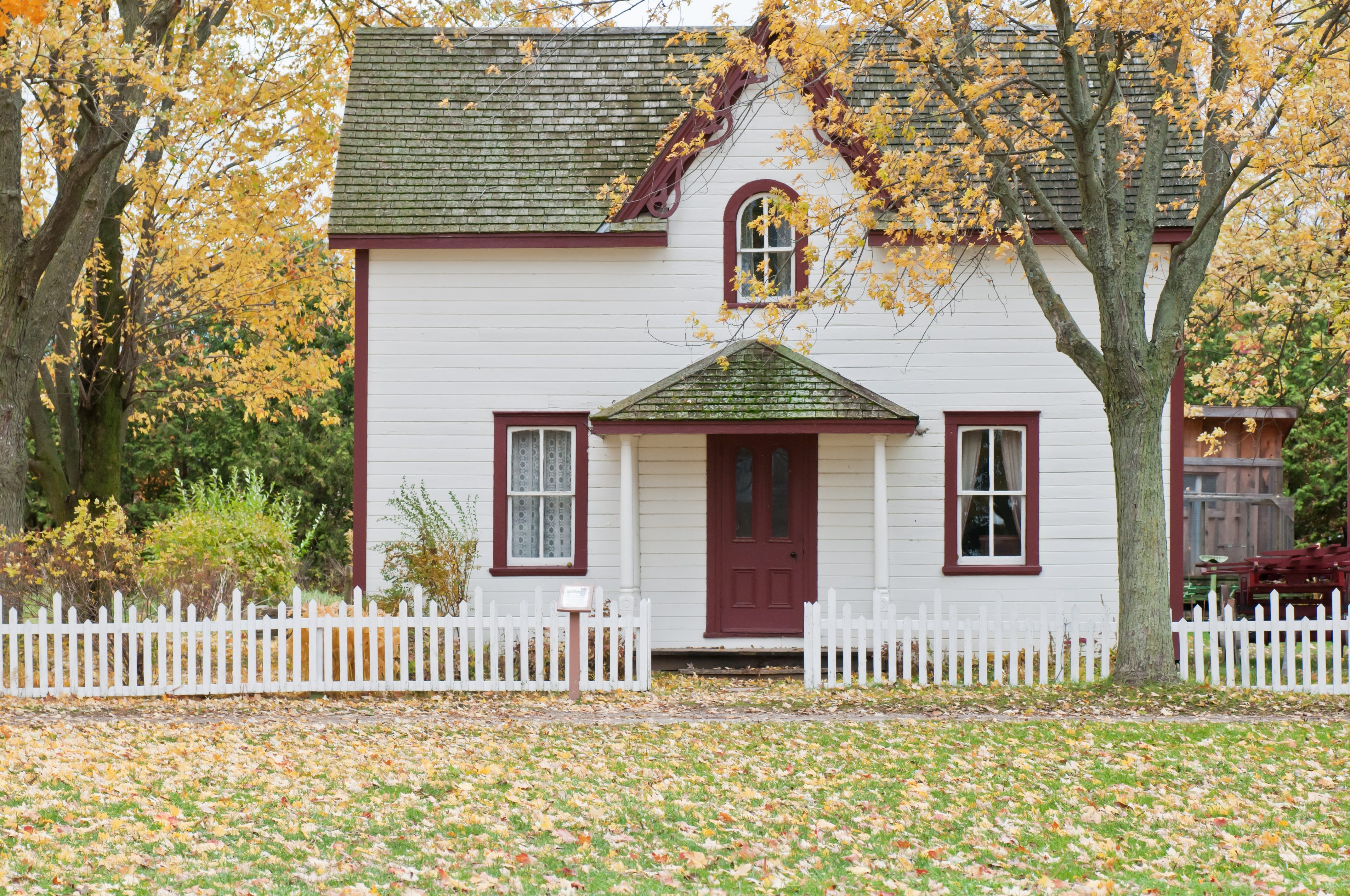 Casa blanca con molduras rojas y una cerca de estacas entre árboles de otoño.