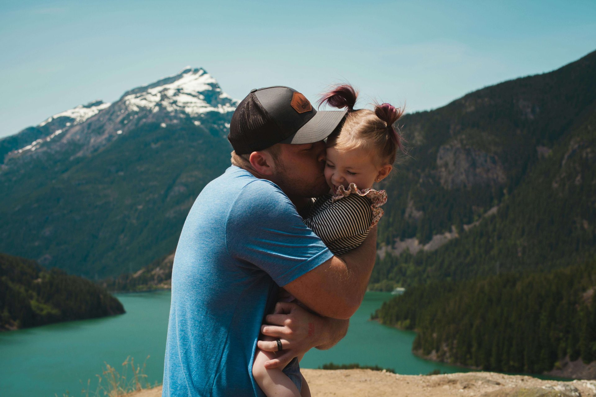 A man kissing a child beside a mountain lake.