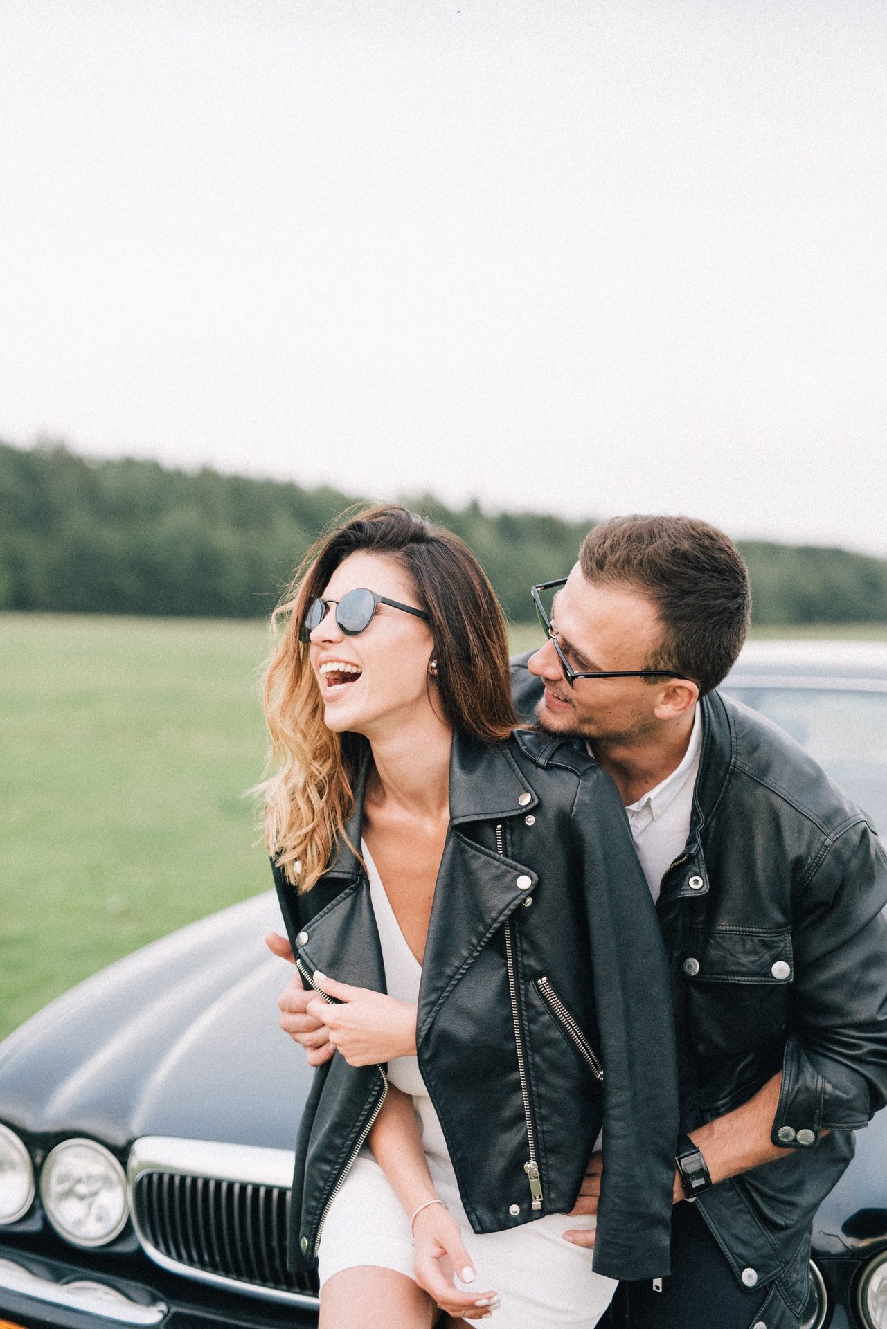 Two adults wearing dark jackets and sunglasses beside a car.