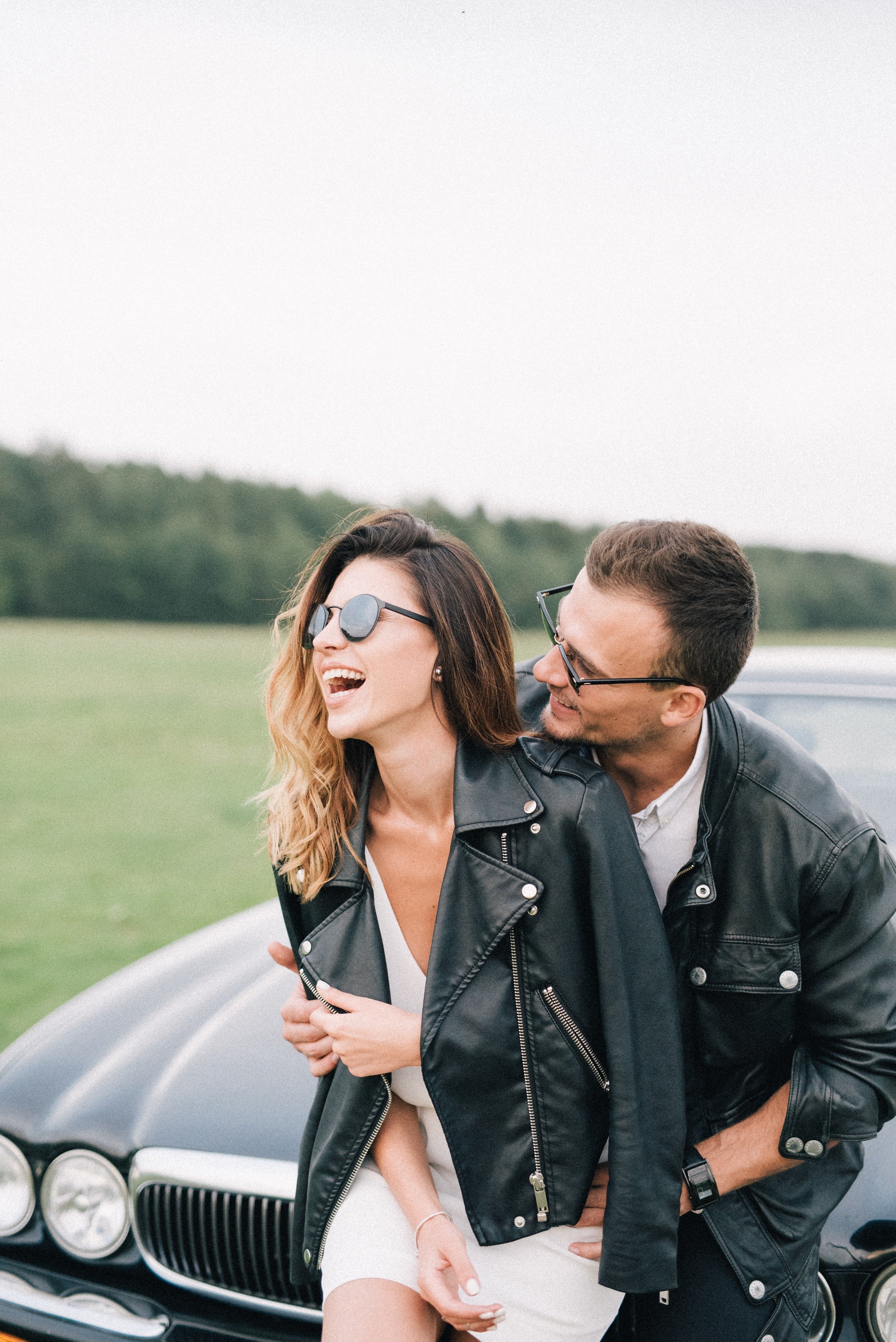 Dos adultos con chaquetas oscuras y gafas de sol junto a un automóvil.