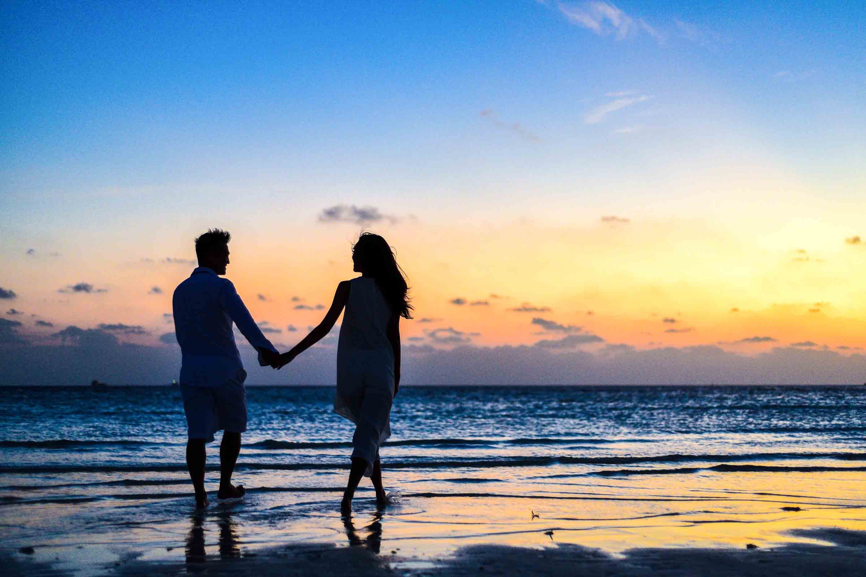 Dos adultos tomados de la mano a la orilla del agua al atardecer.
