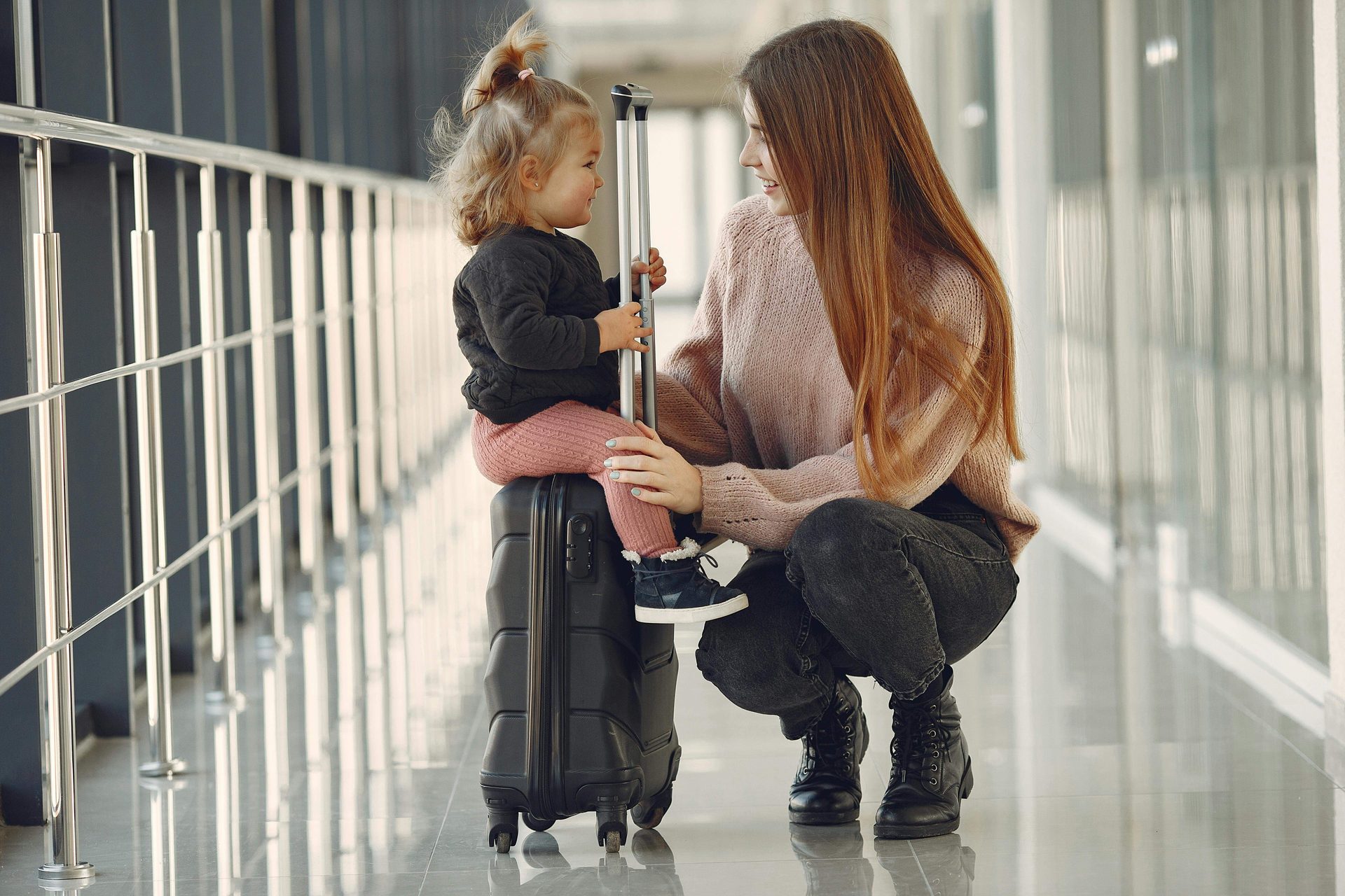 Una mujer junto a una niña sentada sobre una maleta con ruedas.