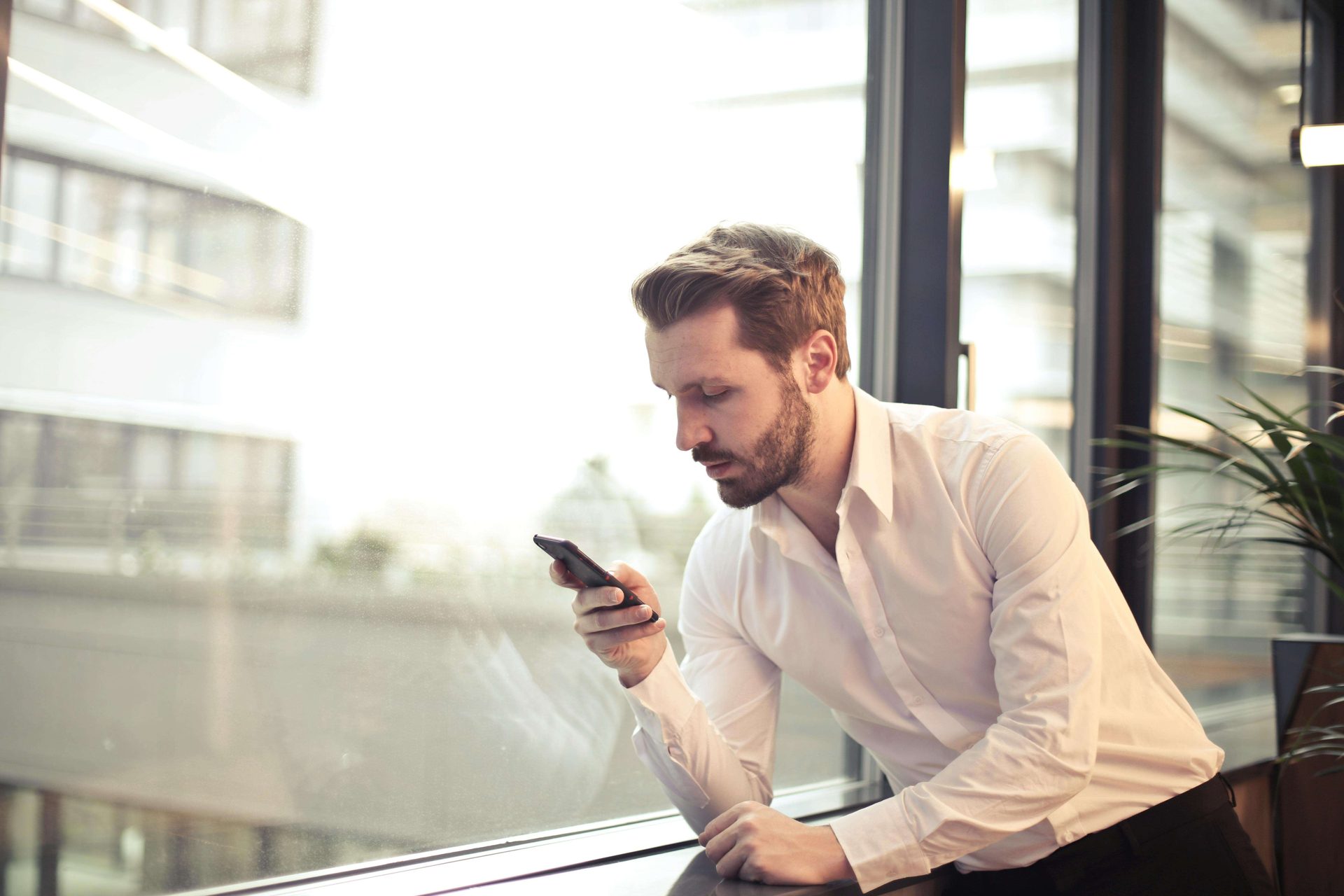 A man looking at his phone beside a window.