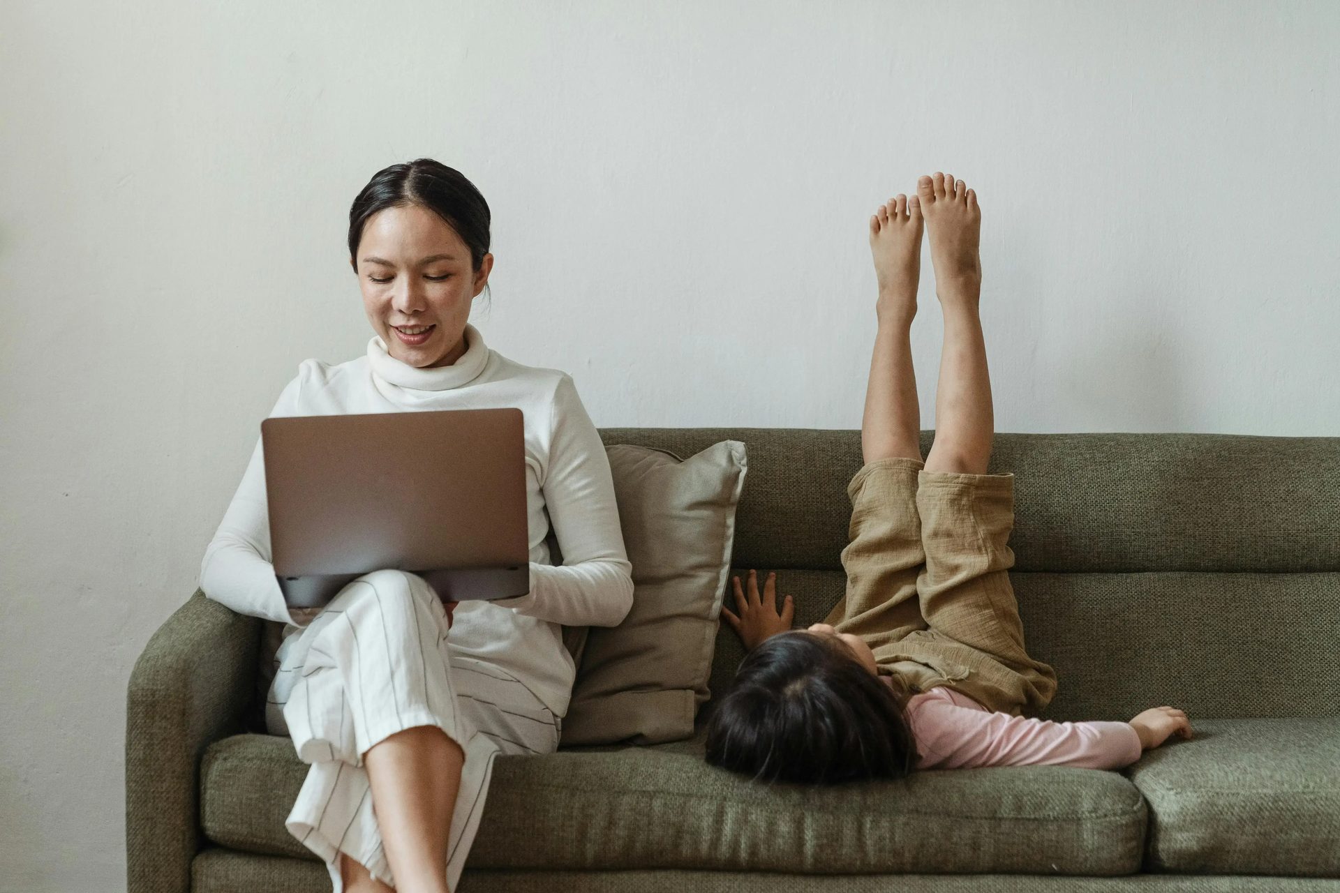 A woman using a laptop while a child plays beside her on a sofa.
