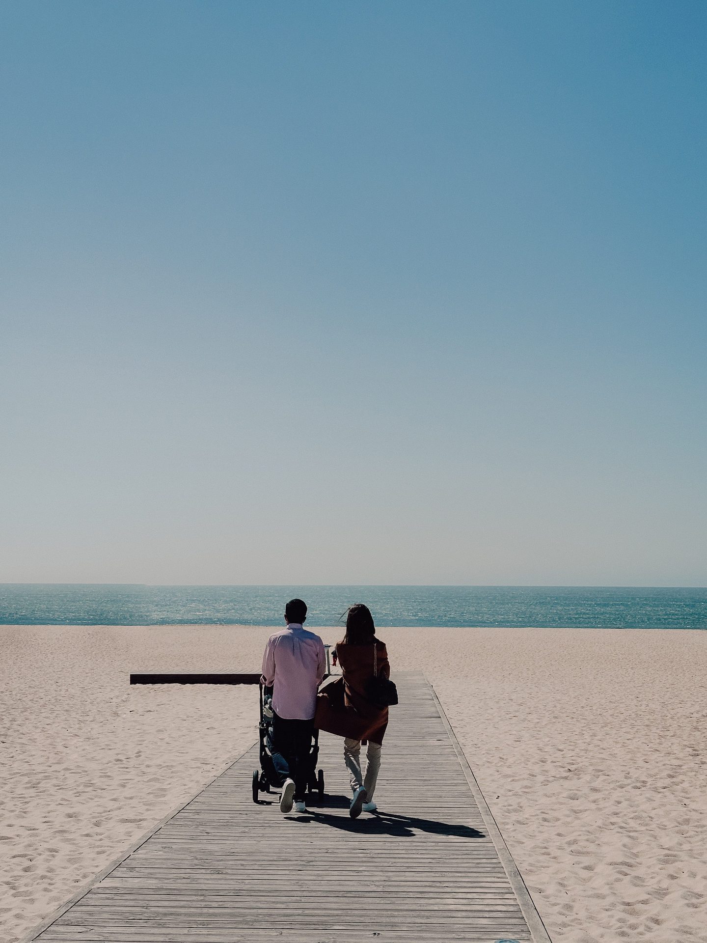 Two adults with a stroller on a wooden path toward a beach.